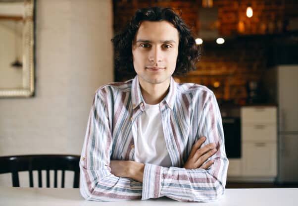 Horizontal Image Of Cool Stylish Young Caucasian Male With Black Curls Relaxing Indoors Sitting At White Desk Keeping Arms Folded, His Look And Posture Expressing Confidence, Smiling At Camera