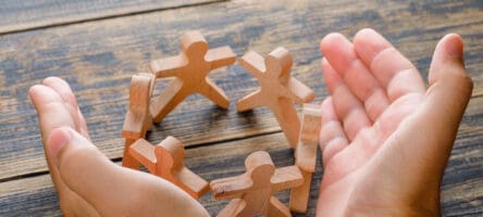 Business Success Concept On Wooden Background High Angle View. Hands Protecting Wooden Figures Of People.