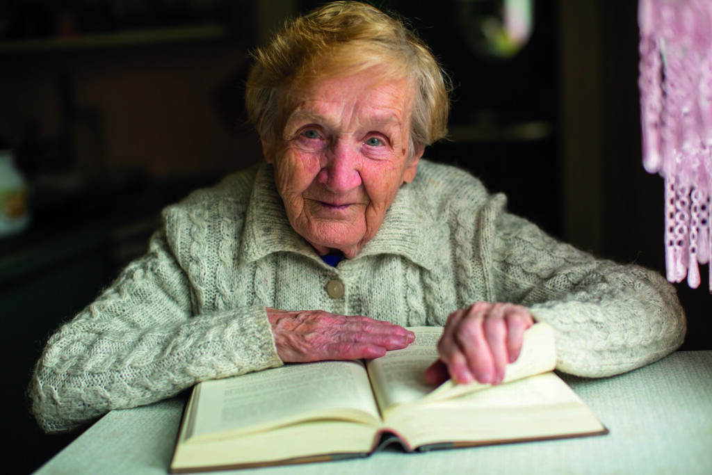 Old Woman Reading A Book Sitting At The Table.