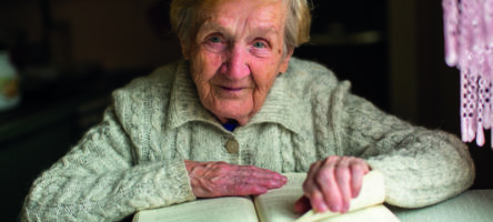 Old Woman Reading A Book Sitting At The Table.