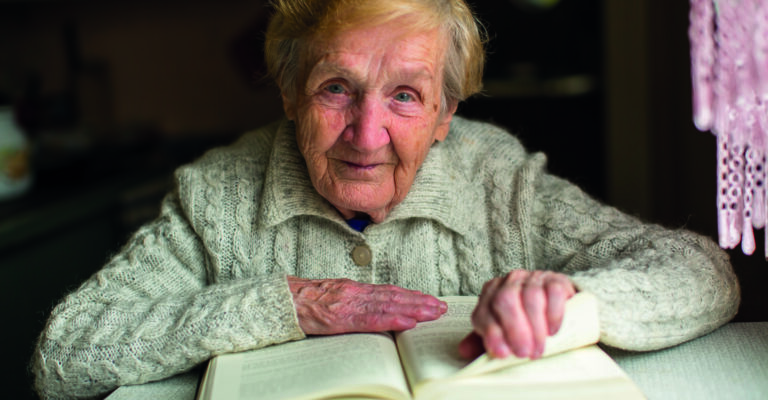 Old Woman Reading A Book Sitting At The Table.