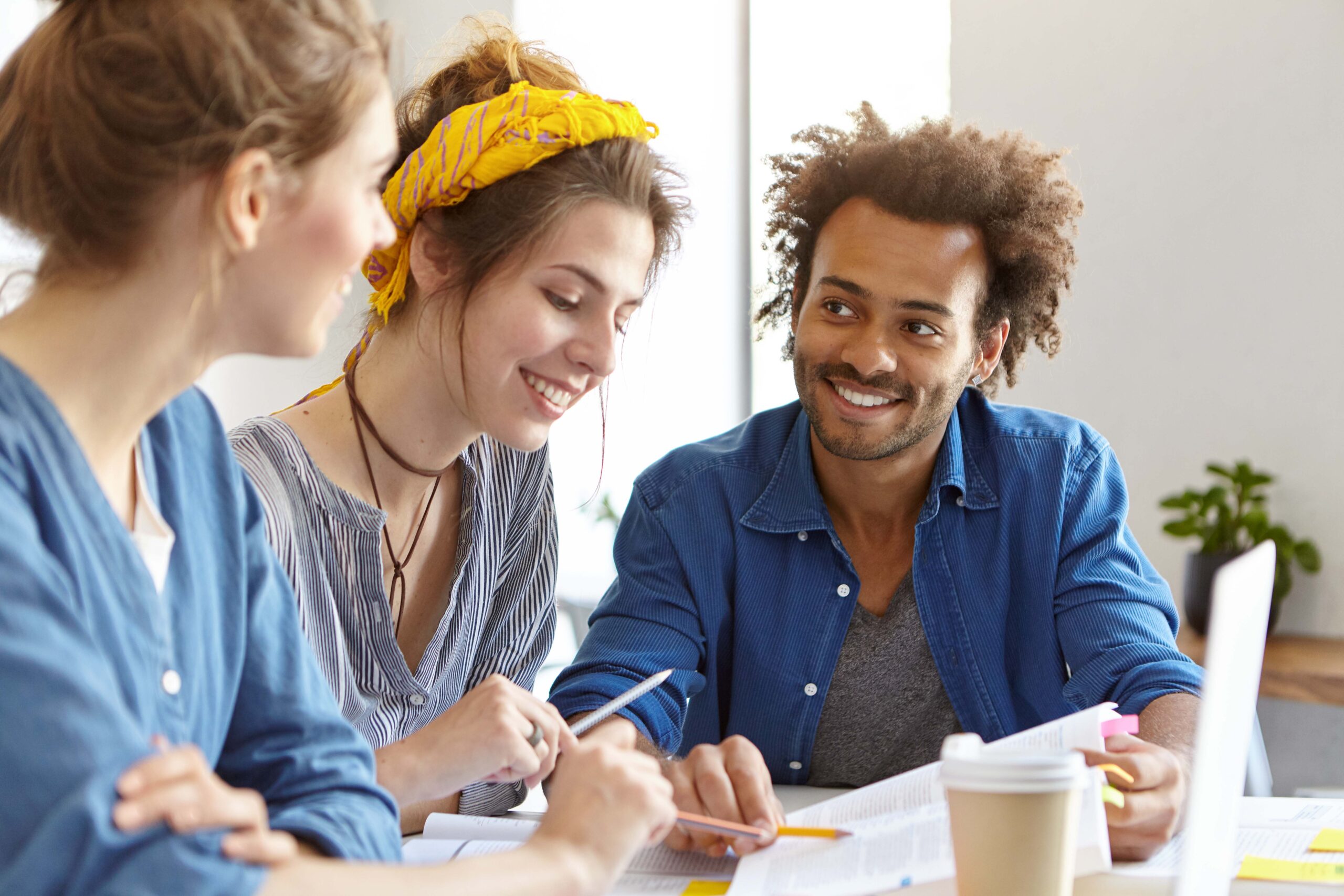 Cheerful Mixed Race People Meeting Together Discussing Main Issues Of Their Work. Multiethnic Friendly Students Looking In Book Pointing At Page With Pencils Doing Their Home Assigment Together